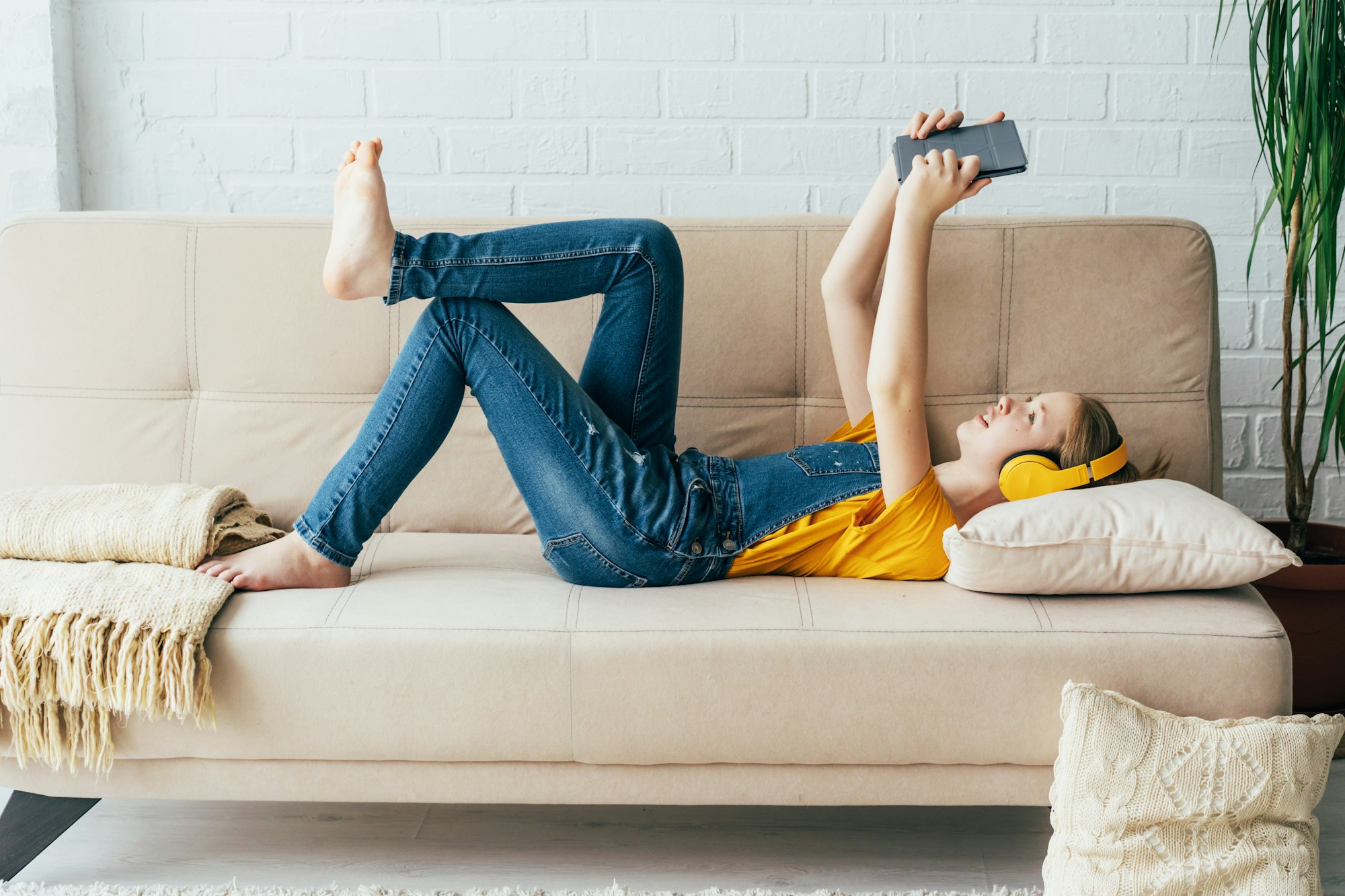 Teenage girl in denim overalls relaxes in pleasure on the couch with an electronic tablet.